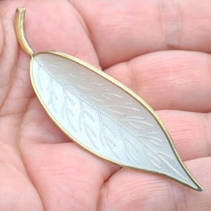 Puede incluir: Broche en forma de hoja de metal dorado con un centro blanco translúcido. El broche presenta delicados detalles de venas dentro de la hoja y un tallo curvo. Los bordes están delineados en dorado, creando un contraste llamativo. Este accesorio de inspiración natural es adecuado para diversas ocasiones.