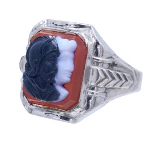 May include: A silver ring featuring a rectangular, reddish-orange cameo with two profiles. One profile is black, the other is white. The ring has a detailed, geometric design on the band.