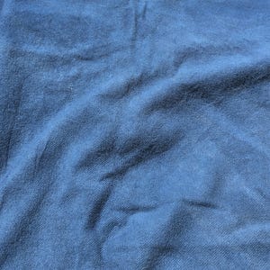 May include: A close-up shot of a textured, blue fabric. The fabric has a slightly wrinkled appearance, with visible folds and subtle variations in the blue hue. The material appears to be soft and possibly a type of knit or woven textile.
