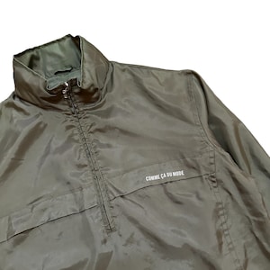 May include: A green, lightweight jacket with a zip closure and a stand-up collar. The jacket has a small pocket on the left chest and the text "COMME CA DU MODE" printed on the right chest.