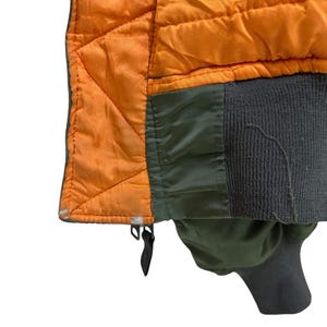 Puede incluir: Primer plano de una chaqueta bomber vintage. La chaqueta presenta un forro naranja acolchado, paneles exteriores verde oliva y un pu&ntilde;o acanalado gris oscuro. El dise&ntilde;o de la chaqueta incluye un patr&oacute;n de costura de diamante en el forro naranja.