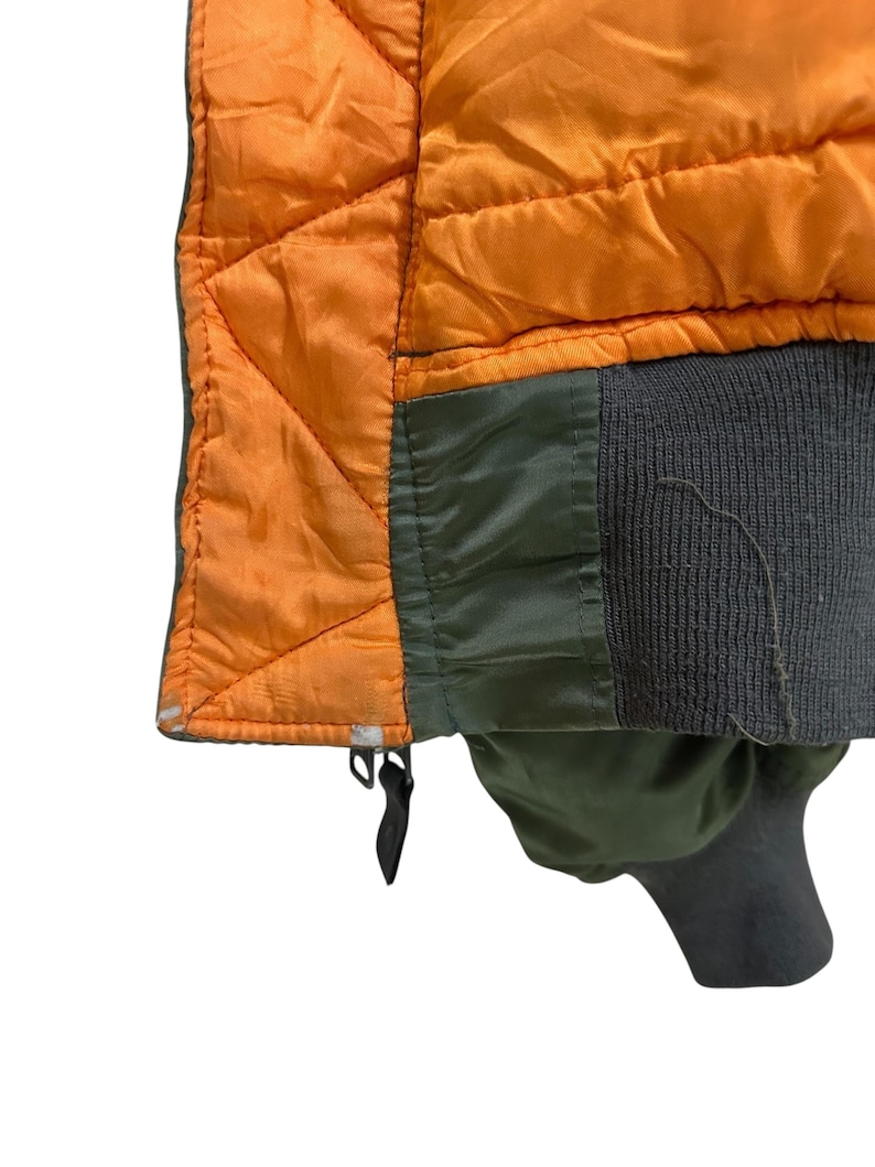 Puede incluir: Primer plano de una chaqueta bomber vintage. La chaqueta presenta un forro naranja acolchado, paneles exteriores verde oliva y un pu&ntilde;o acanalado gris oscuro. El dise&ntilde;o de la chaqueta incluye un patr&oacute;n de costura de diamante en el forro naranja.