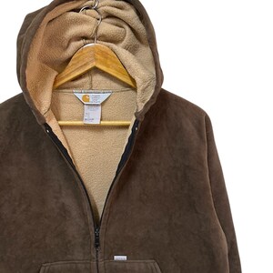 Puede incluir: Una sudadera con capucha de cremallera de color marr&oacute;n con un logotipo de Carhartt en la parte delantera. La sudadera tiene una capucha con forro de polar marr&oacute;n.