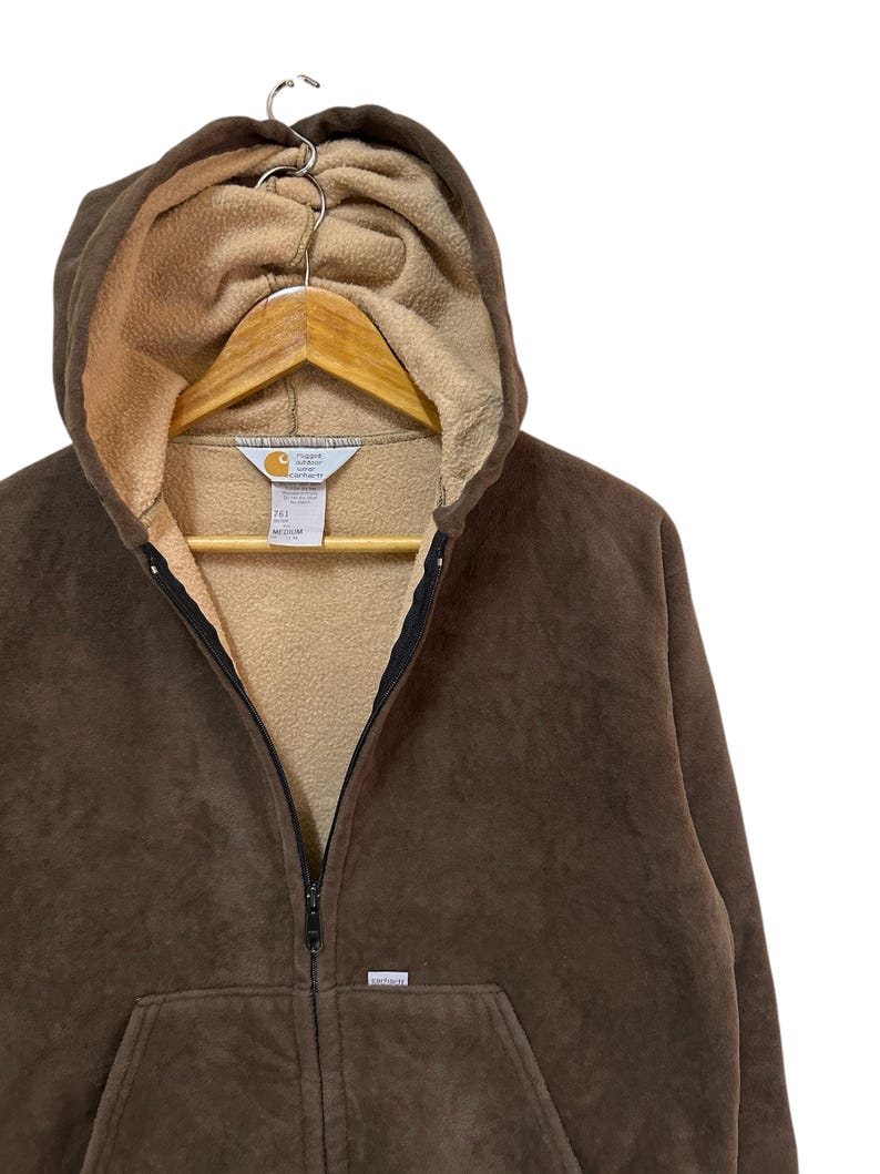 Puede incluir: Una sudadera con capucha de cremallera de color marr&oacute;n con un logotipo de Carhartt en la parte delantera. La sudadera tiene una capucha con forro de polar marr&oacute;n.