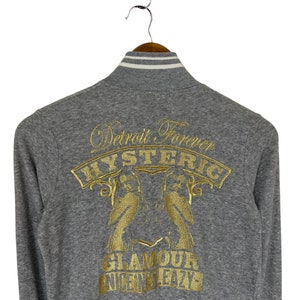 May include: Gray long-sleeve zip-up jacket with a gold embroidered design on the back. The design features two birds, the words "Detroit Forever", "HYSTERIC", "GLAMOUR", and "NICE N' SLEAZY".