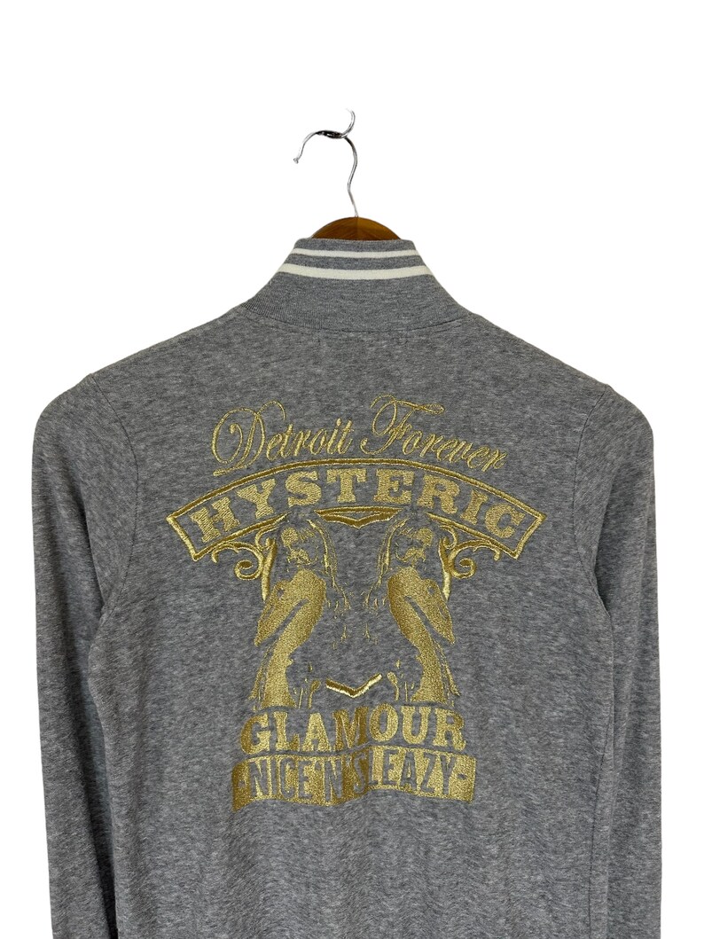 May include: Gray long-sleeve zip-up jacket with a gold embroidered design on the back. The design features two birds, the words "Detroit Forever", "HYSTERIC", "GLAMOUR", and "NICE N' SLEAZY".