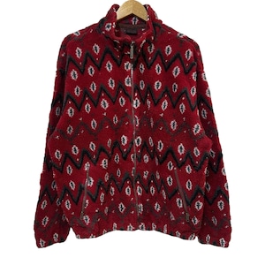 May include: A red fleece jacket with a full zip and a high collar. The jacket has a repeating geometric pattern in black, white, and grey. The jacket is hanging on a wooden hanger.