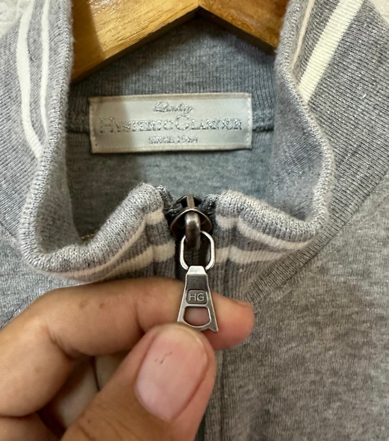 May include: Close-up of a gray zipper on a gray and white striped jacket. The zipper pull has the letters "HG" on it. The label on the jacket reads "Baby Hysteric Glamour Since 1989".