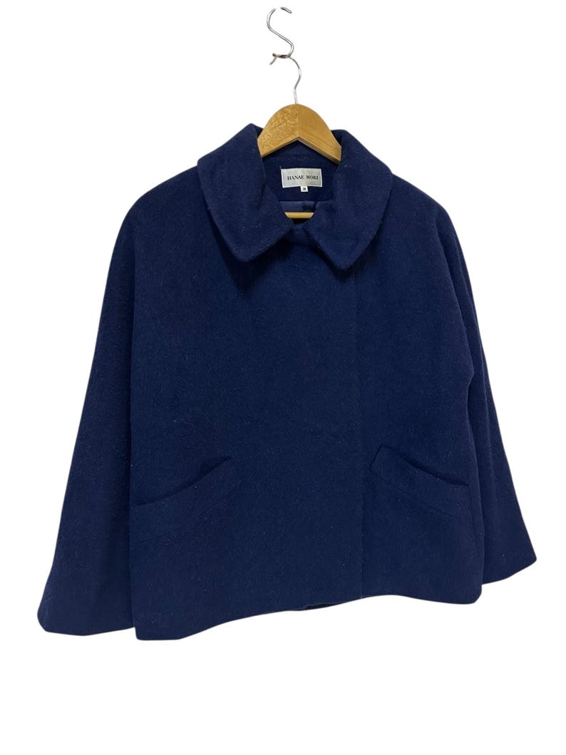 May include: A navy blue wool jacket with a wide collar and two front pockets. The jacket is hanging on a wooden hanger. The brand name "HANAE MORI" is visible on a tag inside the collar.