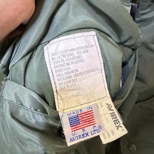 Puede incluir: Primer plano del interior de una chaqueta verde de estilo militar, con una etiqueta que muestra texto como "JACKET, FLYER'S MAN INTERMEDIATE, MA-1" y "AVIREX U.S.A." La etiqueta tambi&eacute;n muestra la bandera estadounidense y las palabras "MADE IN U.S.A."