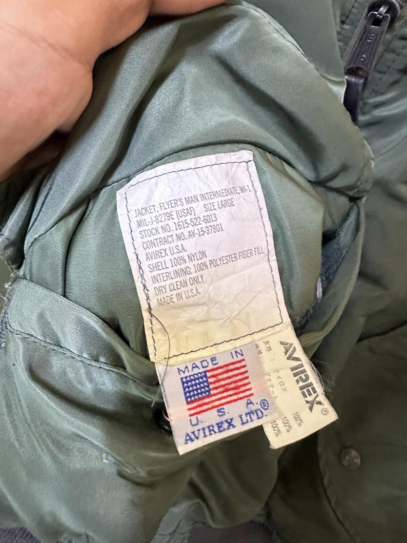 Puede incluir: Primer plano del interior de una chaqueta verde de estilo militar, con una etiqueta que muestra texto como "JACKET, FLYER'S MAN INTERMEDIATE, MA-1" y "AVIREX U.S.A." La etiqueta tambi&eacute;n muestra la bandera estadounidense y las palabras "MADE IN U.S.A."