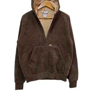Puede incluir: Una sudadera con capucha de cremallera de color marr&oacute;n con un logotipo de Carhartt en la parte delantera. La sudadera tiene una capucha con forro de polar marr&oacute;n.