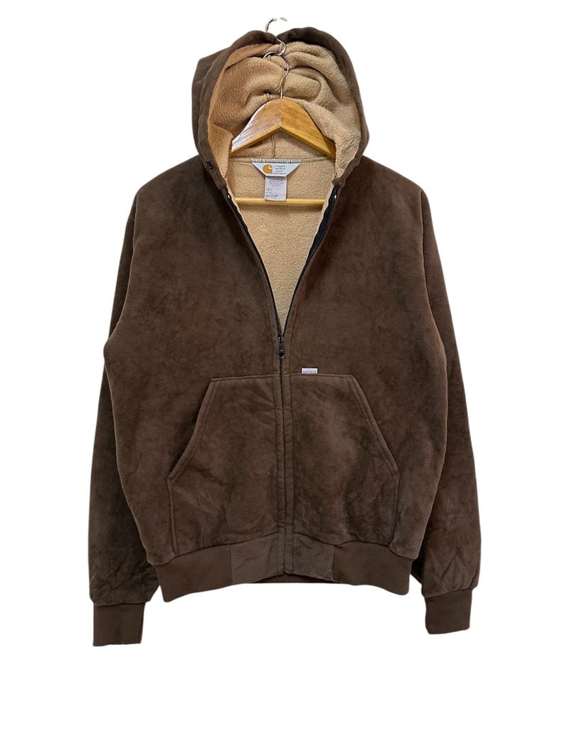 Puede incluir: Una sudadera con capucha de cremallera de color marr&oacute;n con un logotipo de Carhartt en la parte delantera. La sudadera tiene una capucha con forro de polar marr&oacute;n.