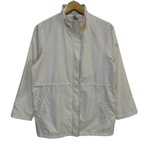 May include: A cream-colored jacket with a high collar and a full-length zipper. The jacket has a white trim along the seams and pockets. It is hanging on a wooden hanger against a white background. The jacket appears to be made of a lightweight material.