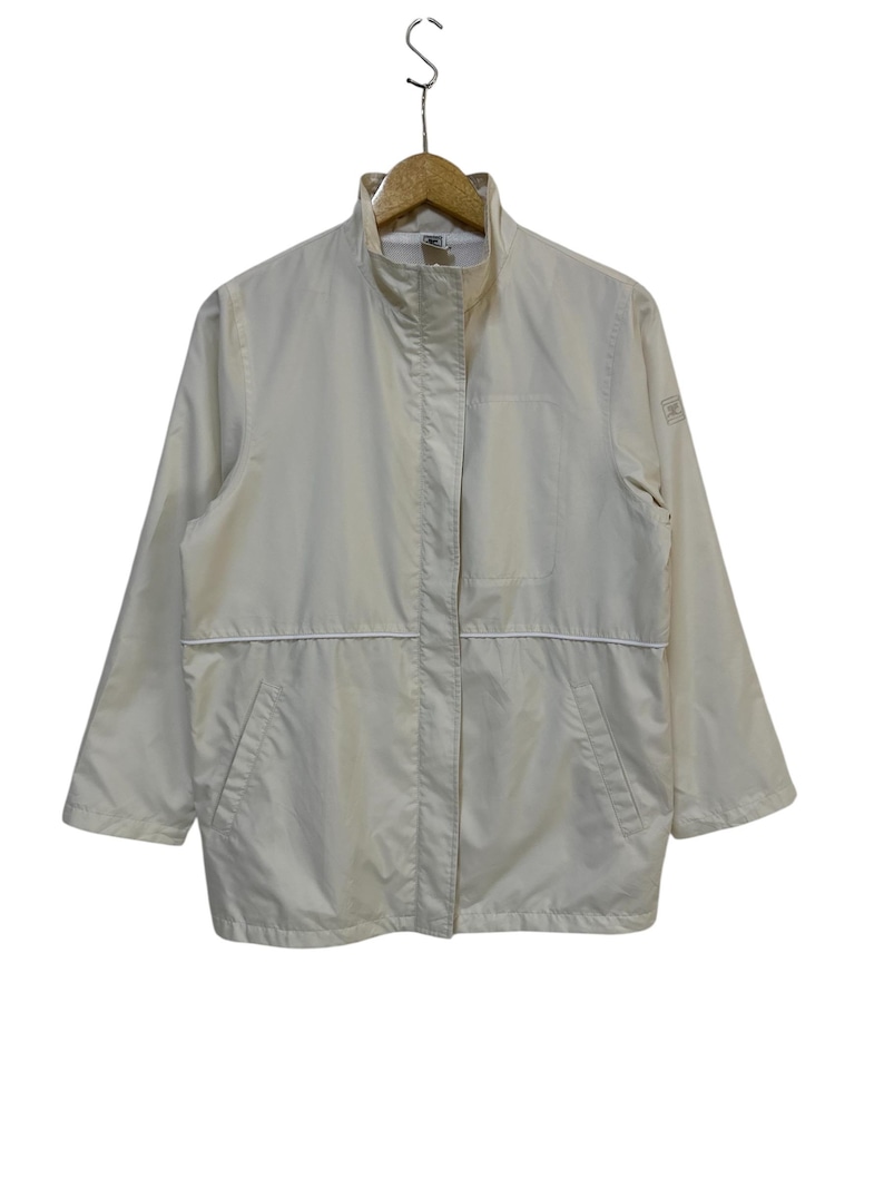 May include: A cream-colored jacket with a high collar and a full-length zipper. The jacket has a white trim along the seams and pockets. It is hanging on a wooden hanger against a white background. The jacket appears to be made of a lightweight material.