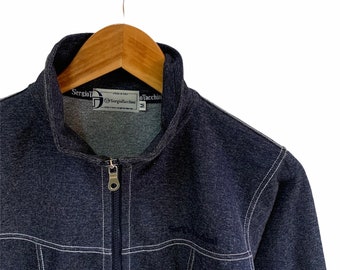 May include: A dark blue zip-up jacket with white stitching. The jacket has a stand-up collar and a zipper closure. The label on the jacket reads "Sergio Tacchini" and "Speed in Style".