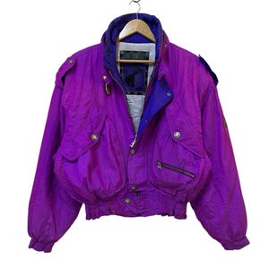 May include: A vintage purple and blue ski jacket with multiple pockets, zippers, and a high collar. The jacket is hanging on a wooden hanger. The jacket has a quilted design and a blue interior lining. The jacket has a retro style.