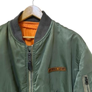 Puede incluir: Chaqueta bomber verde oliva con cuello gris y forro naranja. La chaqueta tiene cierre de cremallera y un peque&ntilde;o parche que dice "AVIREX LTD". Cuelga de una percha de madera.