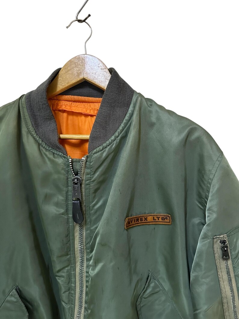 Puede incluir: Chaqueta bomber verde oliva con cuello gris y forro naranja. La chaqueta tiene cierre de cremallera y un peque&ntilde;o parche que dice "AVIREX LTD". Cuelga de una percha de madera.