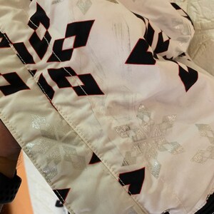 May include: White ski jacket with a black and red geometric pattern and silver snowflake design. The jacket has a zipper closure and a hood.