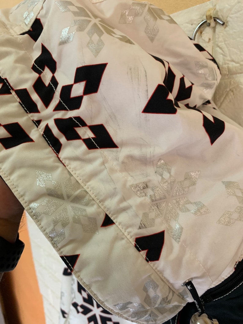 May include: White ski jacket with a black and red geometric pattern and silver snowflake design. The jacket has a zipper closure and a hood.