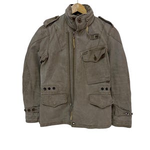 May include: A brown canvas jacket with a zipper closure, multiple pockets, and a high collar. The jacket has a distressed look and features a zippered pocket on the left chest.