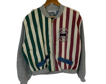 May include: A gray, green, and red striped bomber jacket with a zipper closure. The jacket has a blue denim pocket on each side and a logo patch on the right side that says "Compagnie Americaines".