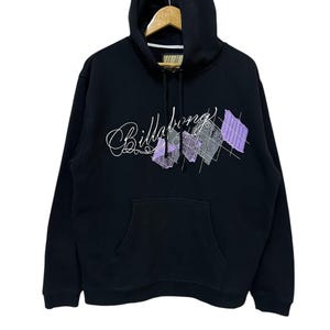 Puede incluir: Sudadera con capucha negra con cordón y bolsillo tipo canguro. En la parte delantera, la palabra "Billabong" en cursiva, con formas geométricas en morado y gris.