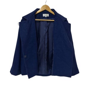May include: A navy blue wool coat with a single button closure and a notched collar. The coat is hanging on a wooden hanger. The interior lining is a darker shade of blue.