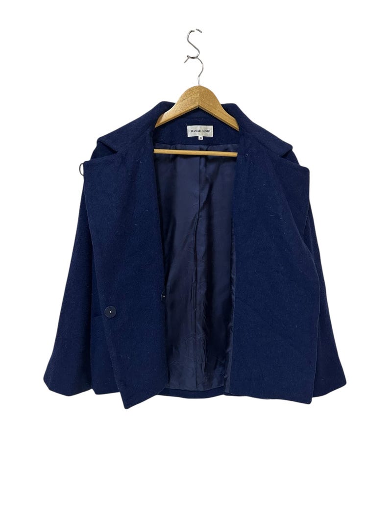 May include: A navy blue wool coat with a single button closure and a notched collar. The coat is hanging on a wooden hanger. The interior lining is a darker shade of blue.