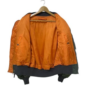 Puede incluir: Chaqueta bomber verde oliva con forro acolchado naranja, colgada de una percha de madera. La chaqueta tiene cuello, pu&ntilde;os y dobladillo de canal&eacute; gris oscuro. La chaqueta est&aacute; desabrochada, revelando el interior.