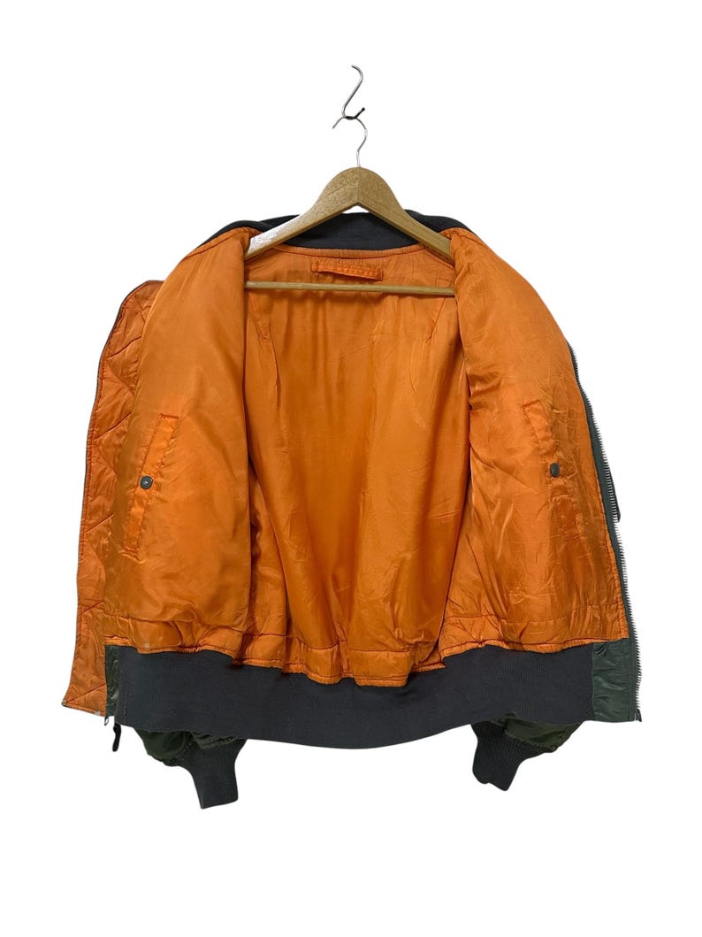 Puede incluir: Chaqueta bomber verde oliva con forro acolchado naranja, colgada de una percha de madera. La chaqueta tiene cuello, pu&ntilde;os y dobladillo de canal&eacute; gris oscuro. La chaqueta est&aacute; desabrochada, revelando el interior.