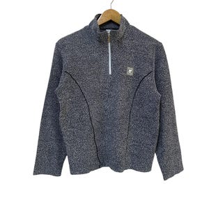 May include: A gray fleece pullover with a half-zip closure and a small logo patch on the chest. The long-sleeved top has a textured appearance and is hanging on a wooden hanger.