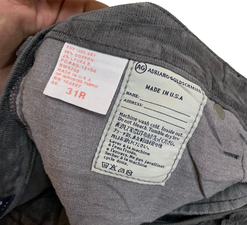 May include: A gray corduroy pant leg with a white care label that reads "Machine wash cold. Inside out. Do not bleach. Tumble dry low. Laver &agrave; la machine &agrave; l'eau froide, &agrave; l'envers. Ne pas javelliser. S&eacute;cher &agrave; la machine cycle doux." The label also includes the brand name "AG Adriano Goldschmied" and "Made in U.S.A."