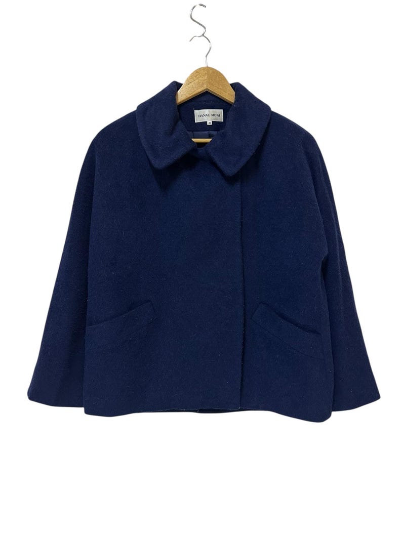 May include: A navy blue wool coat with a wide collar and two front pockets hangs on a wooden hanger. The coat has a simple, classic design and is made of a soft, textured fabric. The brand name "HANAE MORI" is visible on the label.