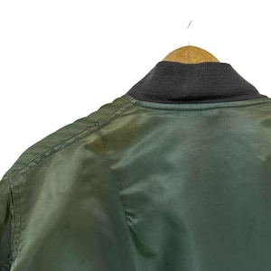 Puede incluir: Primer plano de una chaqueta bomber verde colgada de una percha de madera. La chaqueta tiene un cuello de canal&eacute; gris oscuro y una textura suave y brillante. Los detalles de las costuras son visibles.