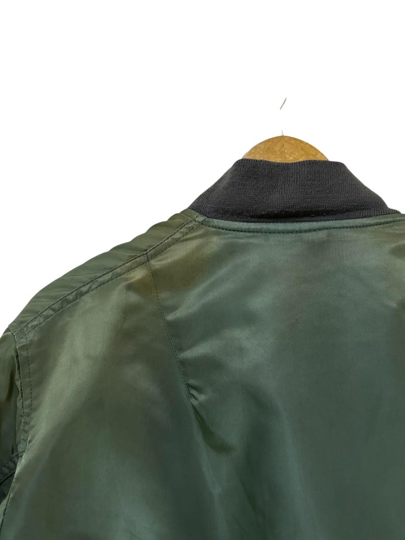 Puede incluir: Primer plano de una chaqueta bomber verde colgada de una percha de madera. La chaqueta tiene un cuello de canal&eacute; gris oscuro y una textura suave y brillante. Los detalles de las costuras son visibles.