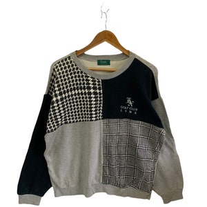 May include: A gray, black, and white patterned sweatshirt with a black and white plaid pattern on the bottom right. The sweatshirt has a green tag that says "Lynx" and a black embroidered logo that says "Golf Club Lynx".