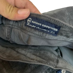 May include: A gray corduroy pair of pants with a blue label that reads "AG Adriano Goldschmied" with a small logo of the letters "AG" in a circle.