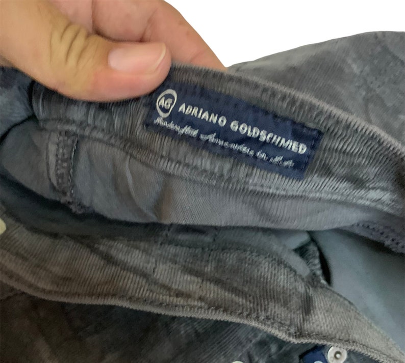 May include: A gray corduroy pair of pants with a blue label that reads "AG Adriano Goldschmied" with a small logo of the letters "AG" in a circle.