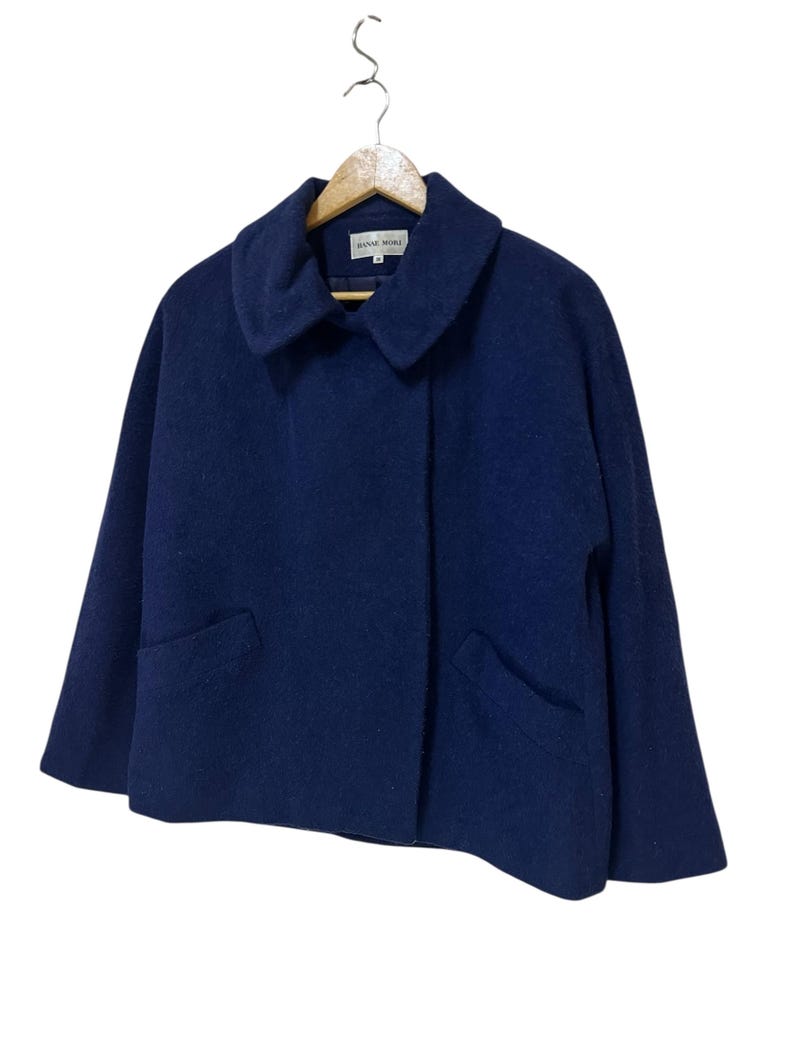 May include: A navy blue wool coat with a wide collar and two front pockets. The coat is hanging on a wooden hanger. The coat is a cropped style.