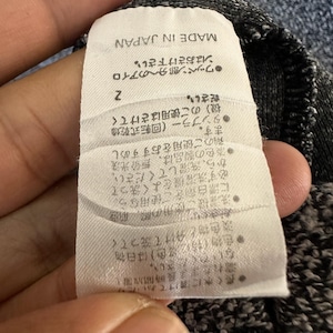 May include: A close-up of a clothing tag with Japanese text and the words "MADE IN JAPAN". The tag is white with black text and is attached to a gray fabric. The tag has care instructions.
