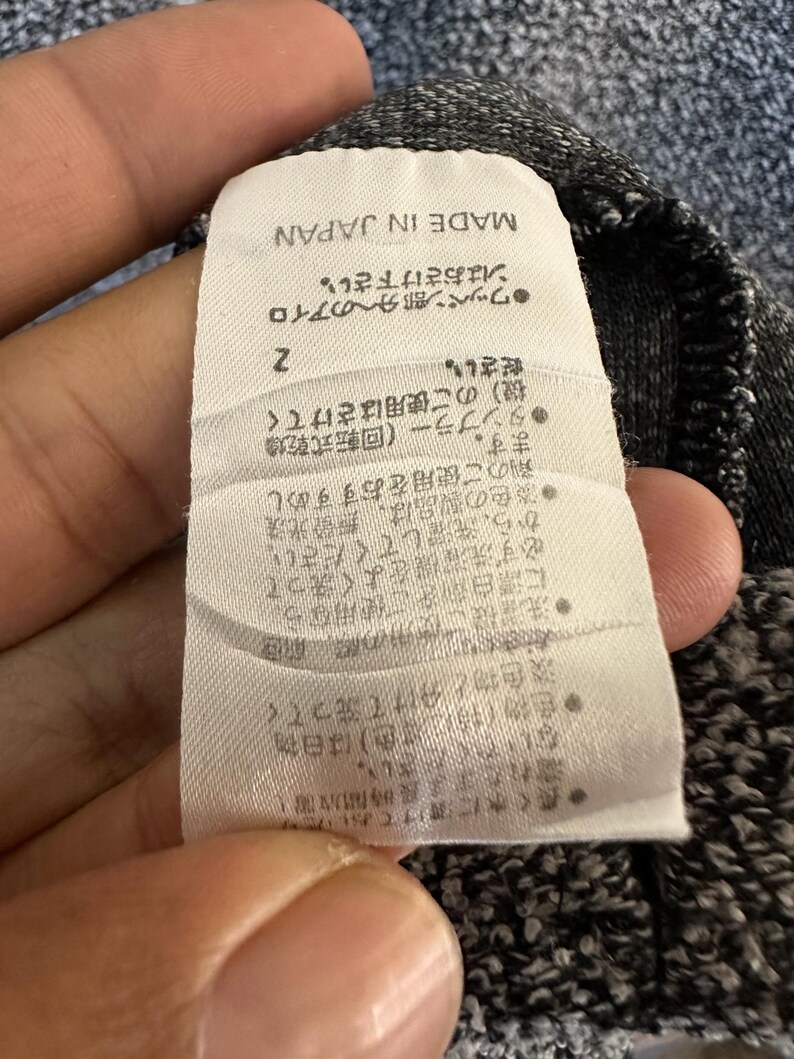May include: A close-up of a clothing tag with Japanese text and the words "MADE IN JAPAN". The tag is white with black text and is attached to a gray fabric. The tag has care instructions.