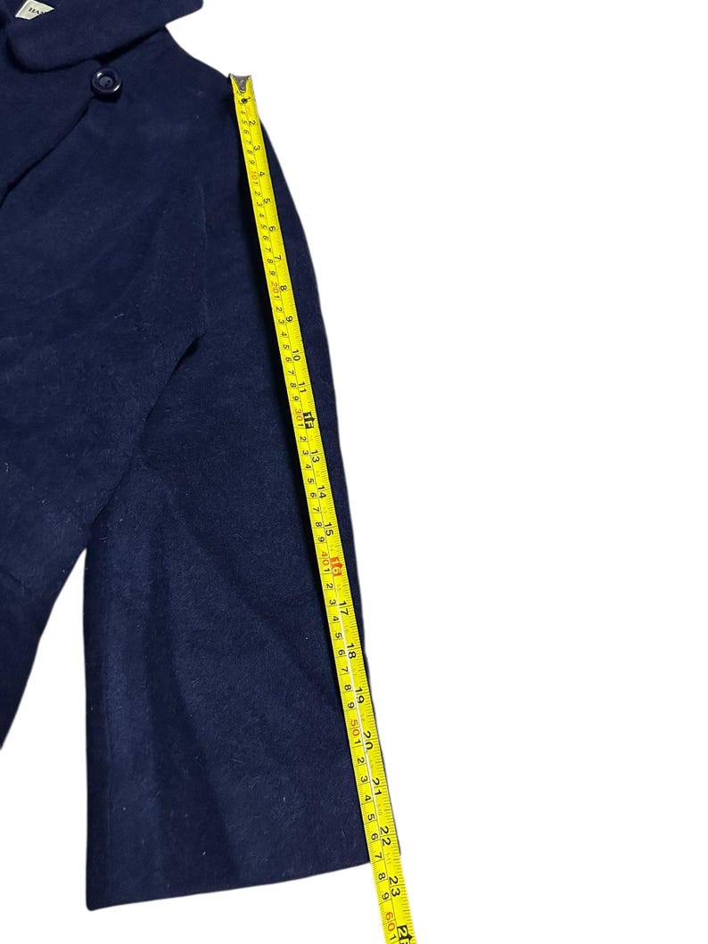 May include: A navy blue wool coat with a button closure and a collar. A yellow measuring tape shows the coat's length. The coat's sleeve is approximately 23 inches long.