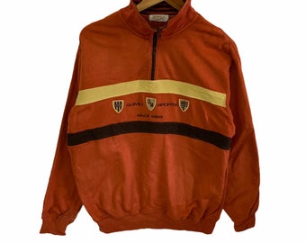 May include: A rust-colored sweatshirt with a black and yellow stripe across the chest. The sweatshirt has a half-zip closure and the text "GUIMVIL SPORT" with a logo and "SINCE 1983" printed on the front.