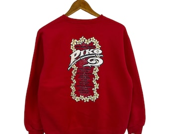 Può includere: Felpa rossa con una stampa grafica bianca della parola "PIKO" circondata da fiori. Il testo "Surf Culture Tradition" e "American Longboard Wear" è anche stampato sulla grafica.