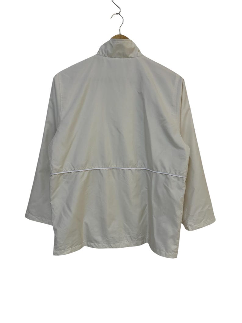 May include: A cream-colored jacket with a stand-up collar and long sleeves. The jacket has a white horizontal stripe across the midsection and is hanging on a wooden hanger. The back of the jacket is visible.