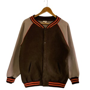 May include: A brown and gray varsity jacket with orange and white stripes on the collar, cuffs, and bottom. The jacket has a button closure and two pockets.