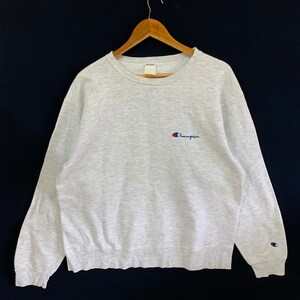 May include: A gray Champion sweatshirt with a small blue and red logo on the front. The logo says "Champion" in a blue font.