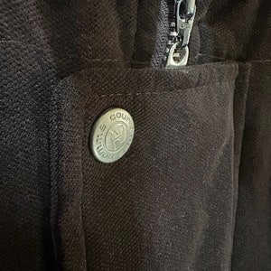 May include: A close-up of a brown zipper on a jacket with a metal button that says "Course" on it.